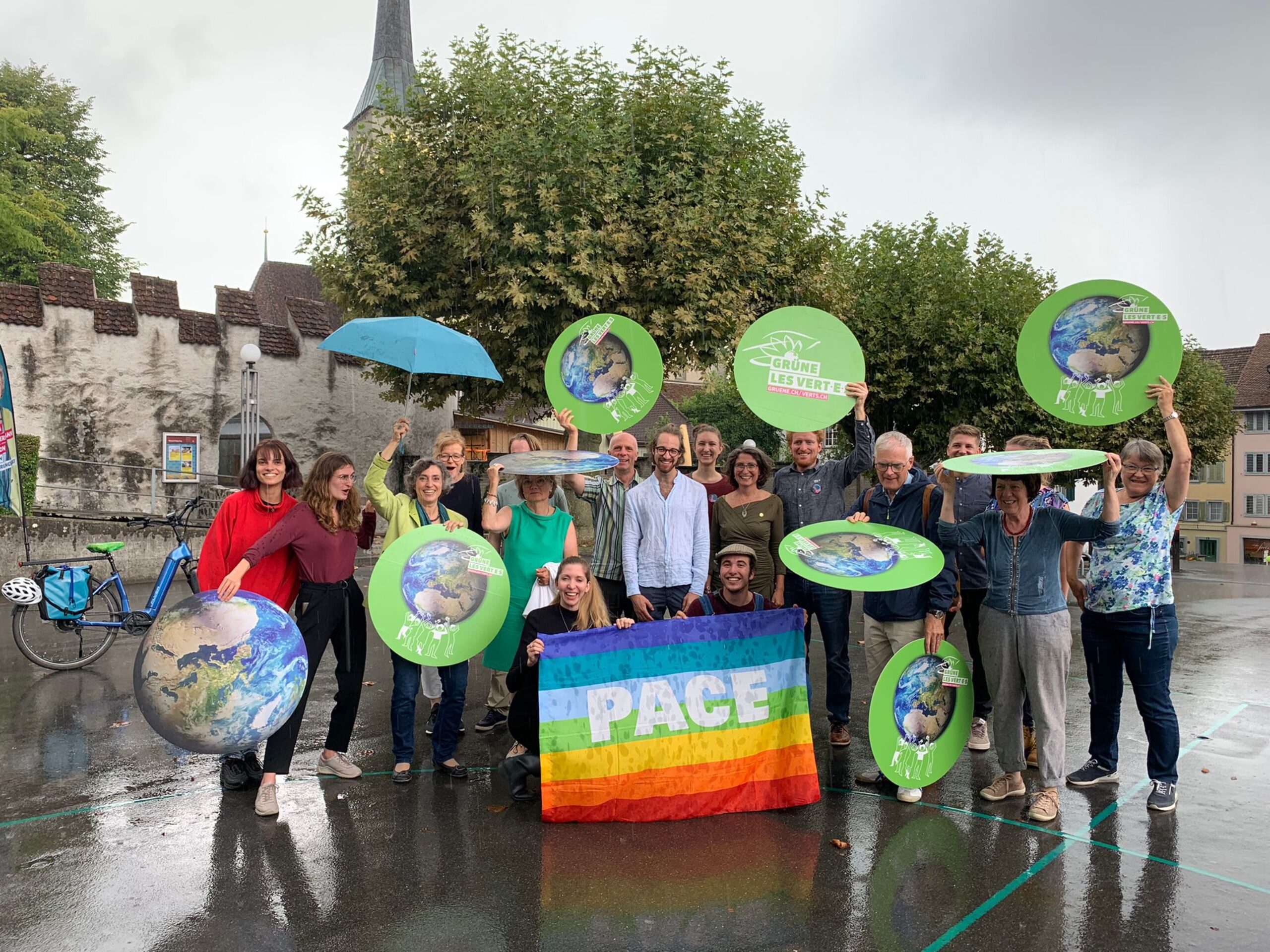 Gruppenfoto der Alternativen - die Grünen. Etwa 20 Helfer*innen und Delegierte welche im Regen stehen. Im Zentrum wird eine Regenbogen-Peace Fahne hochgehalten und überall bunte Kampagnen-Elemnte (Erdkugeln gestämmt von vielen Menschen). Einig nutzen es als Regenschutz. Die Stimmung wirkt trotz Regen sehr fröhlich!