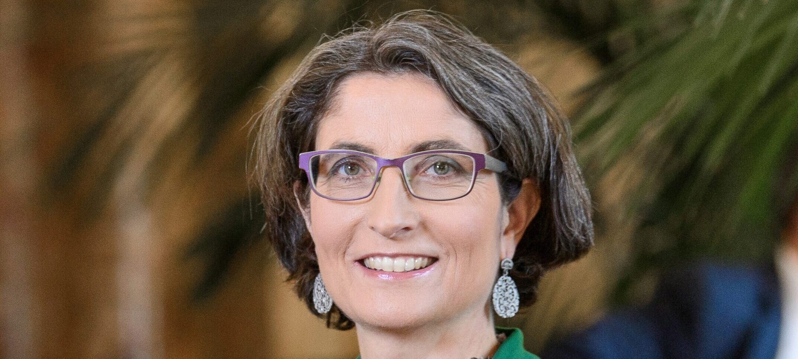 Manuela Weichelt in einem noblen grünen Anzug, einer violetten Brille, gepflegter Frisur, lächelnd, grosse ovale Ohrenhänger bestehend as vielen kleinen Perlen drauf, spannende Kette aus Steinen und weiteren Nautrgegenständen.